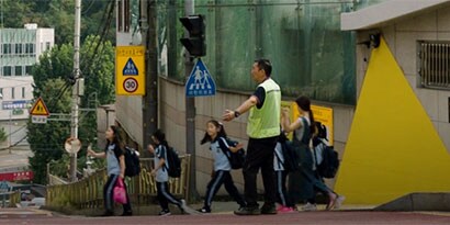 山のある街で、子供たちは蛍光灯の安全ベストを着て大人の指示で通りを渡ります。
