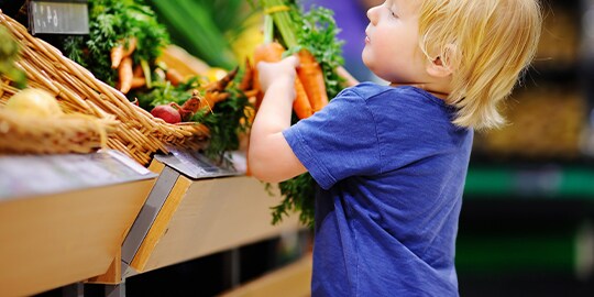 Young child picking up carrots from vegetable display in retail setting.
