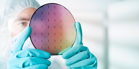 手袋をはめた手でシリコンウエハーを持つクリーンルーム内の人物
