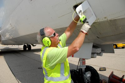 A man wearing a high visibility vest, hearing protection while working on a plane's maintenance panel
