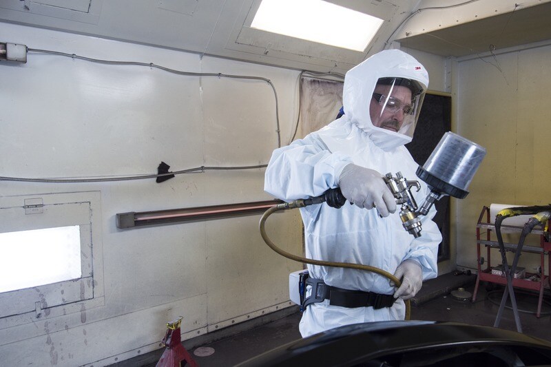 A man wearing a full-body PPE, eye protection, while working on a car with an airbrush
