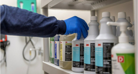 3M の薬剤製品が並ぶ棚を前に、手袋を装着した作業者
