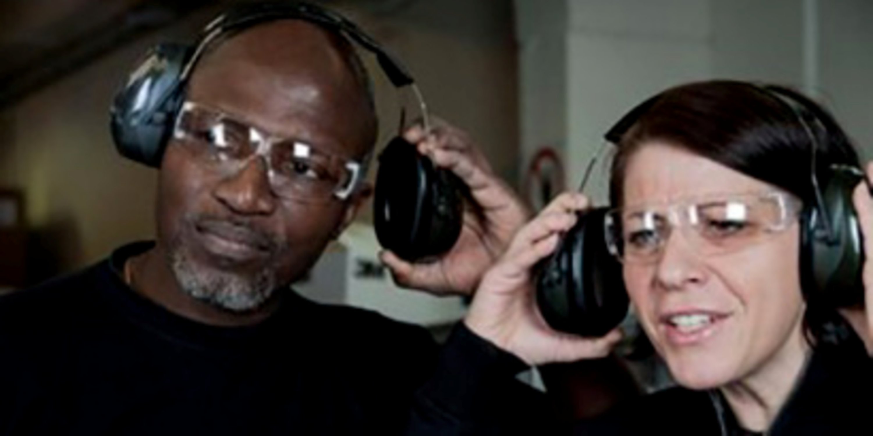 Two people wearing hearing protection, while lifting one of the ear cups to listen to each other
