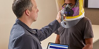 A man checking the fit on a person fully-enclosed in a respirator
