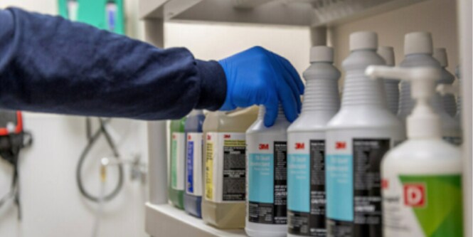 3M の薬剤製品が並ぶ棚を前に、手袋を装着した作業者
