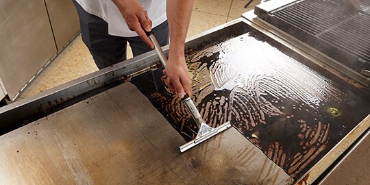 Commercial kitchen worker using 3M Quick Clean griddle cleaning squeegee to clean griddle.

